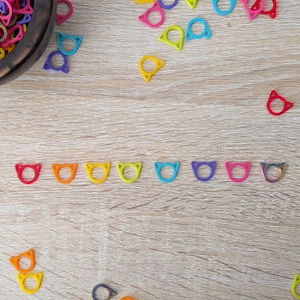 May include: A wooden bowl filled with colourful rubber bands and scattered cat-shaped rings in various colours, including red, orange, yellow, green, blue, purple, pink, and silver. The rings are arranged on a light brown wooden surface.