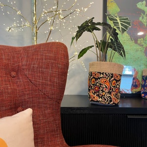 May include: A potted plant in a decorative fabric pot with a black background and a paisley design in red, orange, and yellow. The pot sits on a black surface next to a red upholstered chair.