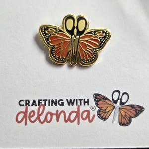 May include: A gold-toned metal enamel pin shaped like a butterfly with orange and black wings and scissor-blade antennae. The pin is attached to a white card featuring the text "CRAFTING WITH delonda" and a matching butterfly scissor graphic.