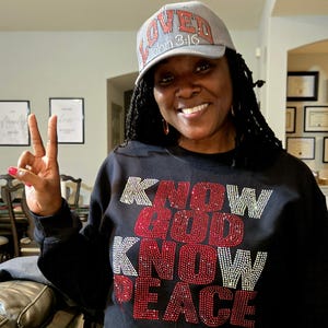 Può includere: Una felpa nera con le parole "KNOW GOD KNOW PEACE" in strass rossi e argentati. È visibile anche un berretto da baseball grigio con la parola "LOVED" e "John 3:16" in strass.