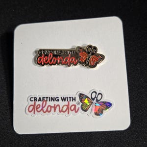 May include: A metal enamel pin and a sticker on a white card. Both feature the text "CRAFTING WITH delonda" and a butterfly with scissors for a body. The pin has orange wings, while the sticker has iridescent, multi-colored butterfly wings.