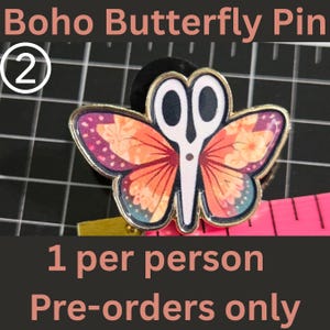 May include: A butterfly-shaped enamel pin featuring a central white scissor design with orange and purple floral patterned wings, outlined in gold metal. A pink measuring tape is visible underneath, showing the pin is approximately 1.5 inches wide.
