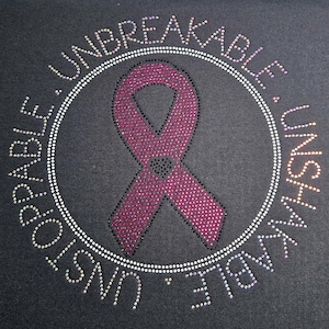 May include: Black fabric with a large, sparkling pink ribbon design, surrounded by a circle of white rhinestones. The words "UNBREAKABLE" and "UNSTOPPABLE" are spelled out in rhinestones around the circle.