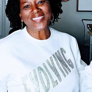 May include: White sweatshirt with the word "EVOLVING" in black and green rhinestones. The person in the image is smiling. A wooden sign that says "Welcome to Flonda's Room" is in the background.