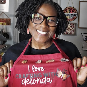 May include: A dark red apron featuring the white text I love CRAFTING WITH delonda and a graphic of a butterfly with scissors as its body. Four small decorative pins are attached to the upper chest area of the fabric, including butterfly designs.