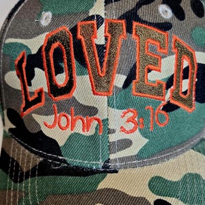 May include: Camouflage baseball cap with the word "LOVED" embroidered in brown with orange trim. The text "John 3:16" is embroidered in orange below the word "LOVED". The cap has a curved brim and a classic six-panel design.