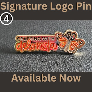 May include: A gold-toned metal pin featuring the text "CRAFTING WITH delonda" in black and red lettering. To the right of the text is a butterfly design with orange and black wings, where the butterfly body is shaped like a pair of open scissors.