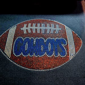 May include: A football design with the word "COWBOYS" in blue rhinestones. The football is brown with white and red rhinestone accents, set against a dark gray background. The design is likely for apparel.