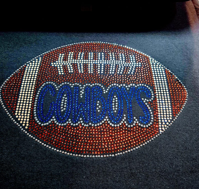 May include: A football design with the word "COWBOYS" in blue rhinestones. The football is brown with white and red rhinestone accents, set against a dark gray background. The design is likely for apparel.