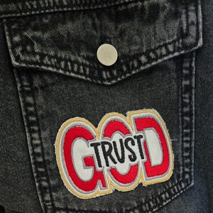 May include: A black denim jacket with a red, white, and yellow patch that says "God Trust" on it.