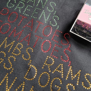 May include: Dark gray t-shirt with rhinestone text that reads "I GOT BEANS GREENS POTATOES TOMATOES LAMBS RAMS DOGS NAME". A package of rhinestones is in the upper right corner.