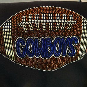 May include: Black garment featuring a football design embellished with rhinestones. The football is brown with white accents and the word "COWBOYS" in blue rhinestones. The design is centered on the garment.