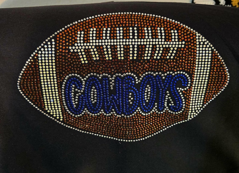 May include: Black garment featuring a football design embellished with rhinestones. The football is brown with white accents and the word "COWBOYS" in blue rhinestones. The design is centered on the garment.