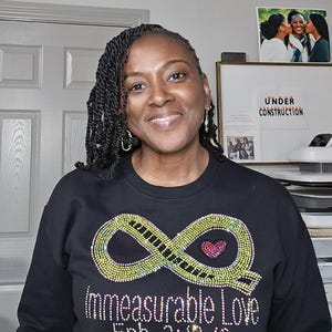 Op de afbeelding: Zwarte sweatshirt met een strass oneindigheidssymbool en een roze hart. De tekst "Immeasurable Love Eph. 3:18-19" staat onder het symbool in strass. De persoon draagt gouden oorringen.