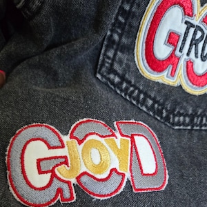Puede incluir: Una chaqueta vaquera con dos parches bordados. El parche superior es rojo, blanco y dorado y dice "G-TRU". El parche inferior es rojo, blanco y dorado y dice "GOOD".