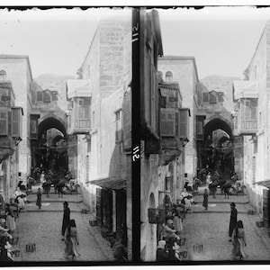 Puede incluir: Una fotografía en blanco y negro de una calle estrecha en una ciudad. La calle está bordeada de edificios que tienen puertas arqueadas y balcones. Hay gente caminando por la calle.