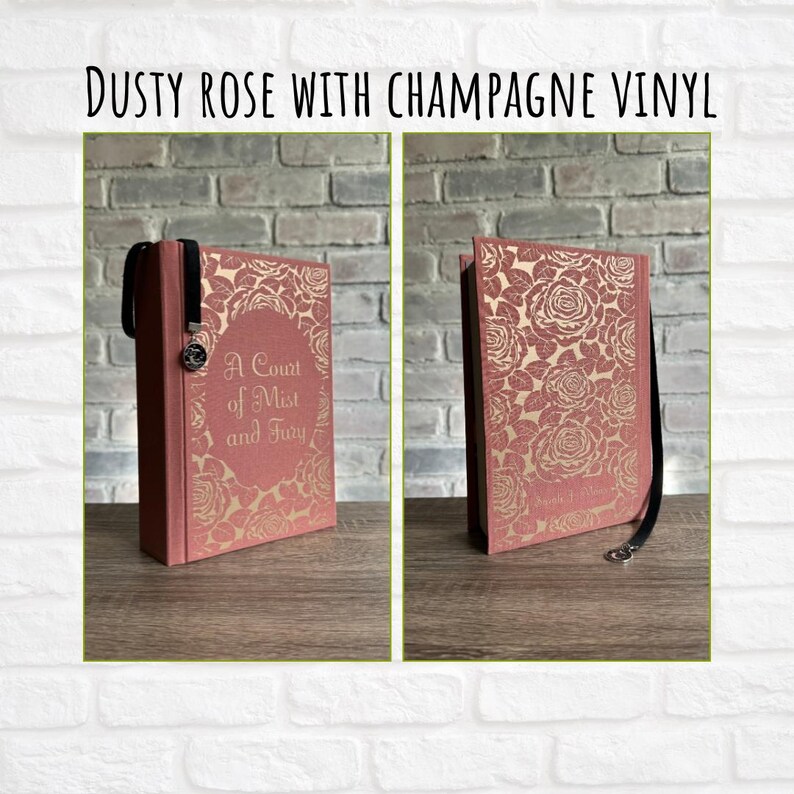 May include: Dusty rose book with champagne vinyl cover featuring a floral rose design. The book has a black ribbon bookmark and the title "A Court of Mist and Fury" is visible. The book is displayed on a wooden surface.