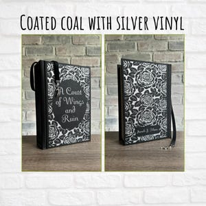 May include: A black book cover with a silver floral design and the title "A Court of Wings and Ruin." The book has a black ribbon bookmark and a silver clasp. The text "Coated Coal with Silver Vinyl" is at the top.