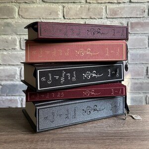 May include: A stack of five hardcover books with decorative floral designs. The books have titles such as "A Court of Thorns and Roses" and "A Court of Silver Flames." The covers are in shades of burgundy, coral, black, and gray.