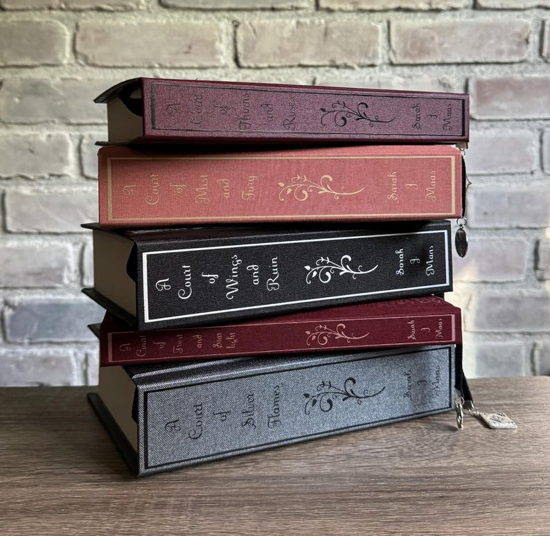 May include: A stack of five hardcover books with decorative floral designs. The books have titles such as "A Court of Thorns and Roses" and "A Court of Silver Flames." The covers are in shades of burgundy, coral, black, and gray.
