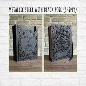 May include: Two views of a book with a metallic steel cover and black foil floral design. The book's title is "A Court of Silver Flames." The cover features a decorative clasp and a black ribbon bookmark.