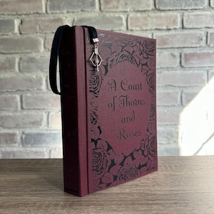 May include: A burgundy book with the title "A Court of Thorns and Roses" in elegant script. The cover features a rose design. A black ribbon bookmark with a silver charm hangs from the top of the book.
