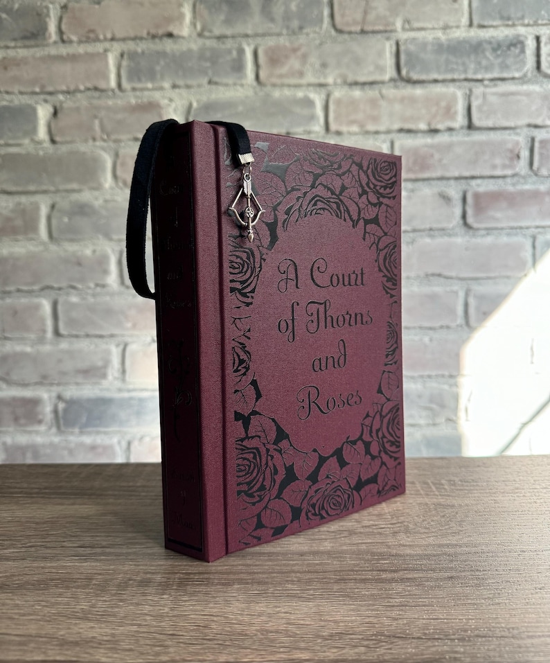 May include: A burgundy book with the title "A Court of Thorns and Roses" in elegant script. The cover features a rose design. A black ribbon bookmark with a silver charm hangs from the top of the book.