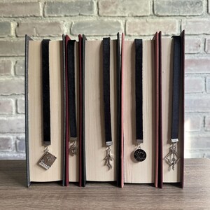 May include: Five hardback books with black velvet ribbon bookmarks. Each bookmark has a different silver charm: a book, a dragon, a feather, a coin, and a decorative arrow. The books are on a wooden surface with a brick background.