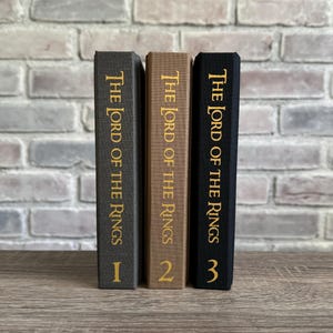 Puede incluir: Tres libros de tapa dura con cubiertas negras, marrones y grises. Los libros se titulan "El Se&ntilde;or de los Anillos" y est&aacute;n numerados del 1 al 3.