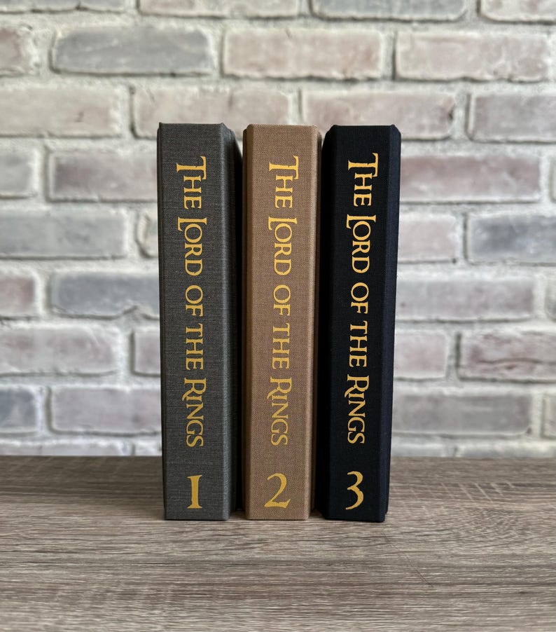 Puede incluir: Tres libros de tapa dura con cubiertas negras, marrones y grises. Los libros se titulan "El Se&ntilde;or de los Anillos" y est&aacute;n numerados del 1 al 3.