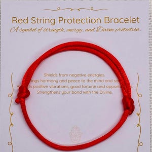 May include: A red string protection bracelet, symbolising strength and protection, is shown on a white card inside a white pouch. The card has text that reads "Red String Protection Bracelet". The bracelet is made of a red string.