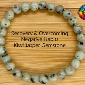 May include: A bracelet made of light green and white kiwi jasper gemstone beads. The bracelet is on a wooden surface. The text "Recovery & Overcoming Negative Habits Kiwi Jasper Gemstone" is written on the image.