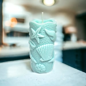 May include: A light blue candle with a cylindrical shape, adorned with seashell and starfish designs. The candle has a textured surface and a white wick. The candle is a decorative item with a coastal theme.