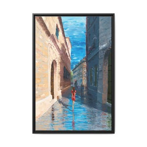 May include: Oil painting of a European street scene with a woman in a red dress walking down a wet cobblestone street. The buildings are painted in shades of beige and blue, with a bright blue sky above. The artwork is framed in a black frame.