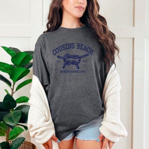 May include: A dark charcoal grey short-sleeve t-shirt featuring a navy blue graphic of a nautical rope knot. The text reads COUSINS BEACH, EST 2021, and NORTH CAROLINA. The shirt is worn with a cream-coloured knit cardigan and light blue denim shorts.