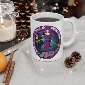 Puede incluir: Taza de cerámica blanca con un gráfico morado y azul de una mujer sosteniendo un bastón. El gráfico incluye el texto "NROL-35 VANDENBERG ATLAS V".