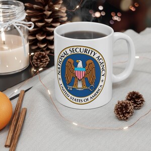 Puede incluir: Una taza de cerámica blanca con el sello de la National Security Agency en la parte frontal. El sello muestra un águila calva con un escudo en el pecho, sosteniendo una llave en sus garras, rodeado por el texto National Security Agency.