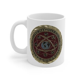 Puede incluir: Taza de cerámica blanca con un diseño dorado y rojo. El diseño presenta un círculo con las palabras "National Security Operations Center" y un globo rodeado de símbolos.