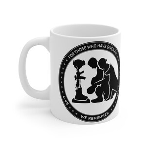 May include: White ceramic coffee mug with a black silhouette of a soldier kneeling with a dog and a fallen soldier memorial. The text around the image reads "For those who have given all, L-45, We Remember".