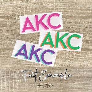 May include: Three white stickers with the letters "AKC" in different colors. The first sticker has pink letters, the second has green letters, and the third has purple letters. The stickers are on a wood background. The text "Font Example" and "Ari Kate Co" are written below the stickers.