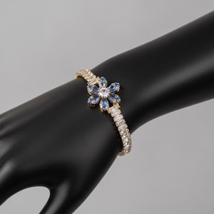 Puede incluir: Una pulsera dorada con una pieza central en forma de flor. El diseño floral presenta cristales azules y transparentes. La pulsera está engastada con cristales transparentes. La joya se exhibe en una mano de maniquí negra.