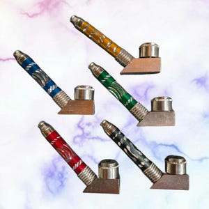 May include: Five metal pipes with colorful, patterned stems in blue, yellow, green, red, and black. Each pipe has a silver-colored bowl and a rectangular base. The pipes are arranged at different angles against a soft, blurred background.