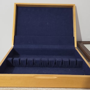 May include: A rectangular wooden storage box with a light brown finish and a hinged lid. The interior is lined with dark blue velvet fabric and features a moulded insert with twelve individual slots designed for organising flatware or utensils.