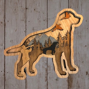 May include: A layered wooden art piece in the shape of a dog, featuring a hunting scene. The design includes a hunter, mountains, trees, and flying birds, all within the dog's silhouette. The piece uses natural wood tones and a dark dog silhouette.