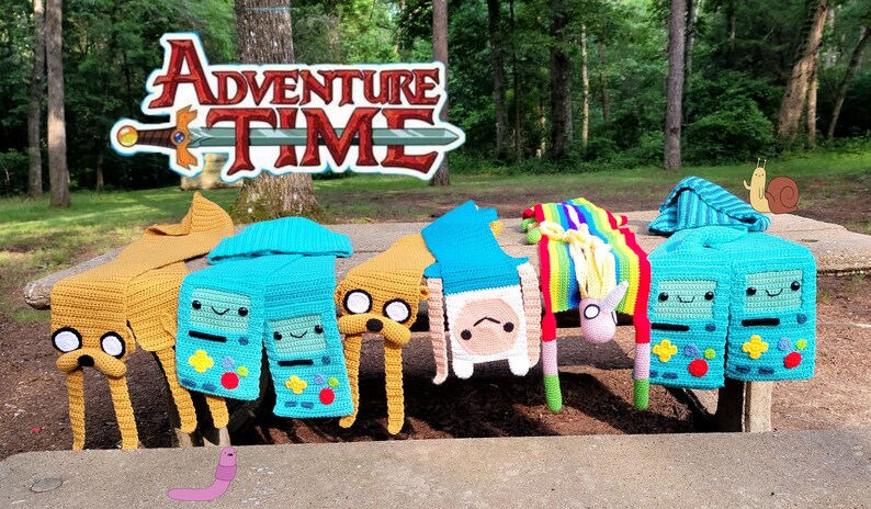 以下が含まれることがあります： フィン・ザ・ヒューマン、ジェイク・ザ・ドッグ、BMO、ユニコーンなど、アドベンチャータイムのキャラクターのモチーフのクロシェ編みスカーフ。スカーフは、アドベンチャータイムのロゴが背景にある木製ピクニックテーブルに飾られています。