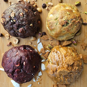 May include: Four different types of cookies on a wooden cutting board. The cookies are a chocolate cookie with hazelnuts, a green cookie with pistachios, a red cookie with white chocolate chips, and a brown cookie with caramel and cookie crumbs.