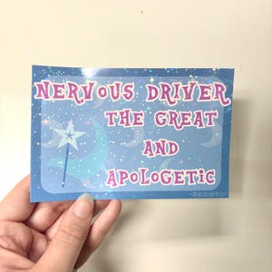 Puede incluir: Una pegatina rectangular con fondo hologr&aacute;fico azul que muestra estrellas, lunas y una varita con punta de estrella. El texto dice: NERVOUS DRIVER THE GREAT AND APOLOGETIC. Las letras son rosas con borde blanco sobre un patr&oacute;n celestial.