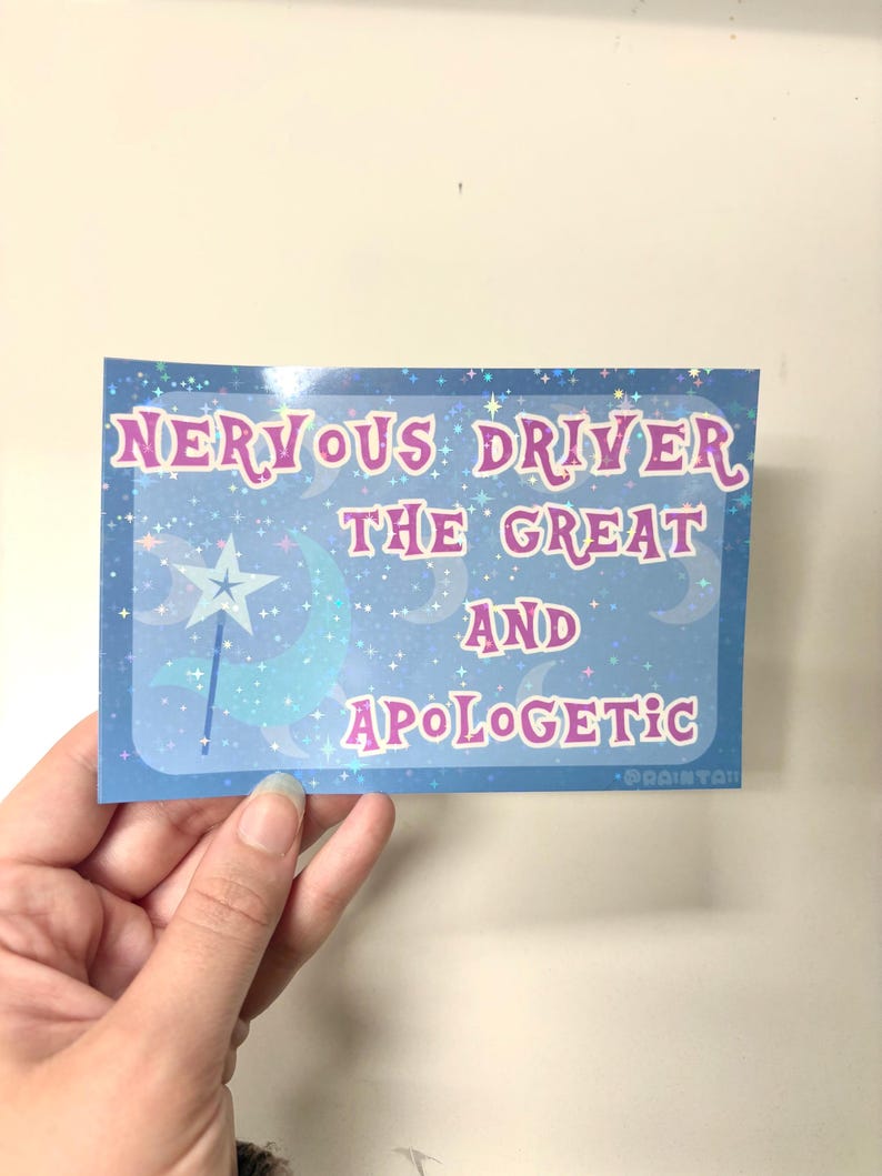 Puede incluir: Una pegatina rectangular con fondo hologr&aacute;fico azul que muestra estrellas, lunas y una varita con punta de estrella. El texto dice: NERVOUS DRIVER THE GREAT AND APOLOGETIC. Las letras son rosas con borde blanco sobre un patr&oacute;n celestial.
