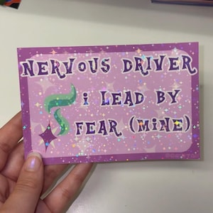 Puede incluir: Una pegatina rectangular con acabado hologr&aacute;fico, fondo morado claro y patrones de estrellas blancas. El texto dice NERVOUS DRIVER I LEAD BY FEAR (MINE) en letras moradas oscuras, junto a un gr&aacute;fico de estrella verde y morado.