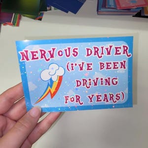 Pu&ograve; includere: Un adesivo rettangolare con sfondo azzurro, nuvole bianche e un motivo a stelle olografiche. Il testo NERVOUS DRIVER (I'VE BEEN DRIVING FOR YEARS) &egrave; scritto in rosso, accompagnato da una nuvola bianca con un fulmine a forma di arcobaleno.